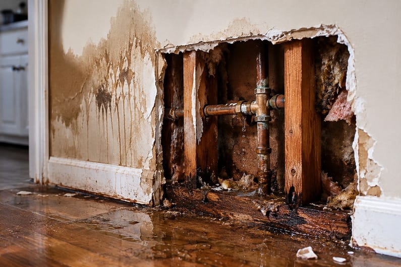 Hidden plumbing leak behind damaged wall with exposed copper pipes, water stains, and floor moisture inside a home