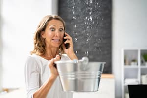 Woman calling plumber while catching water from ceiling leak in bucket